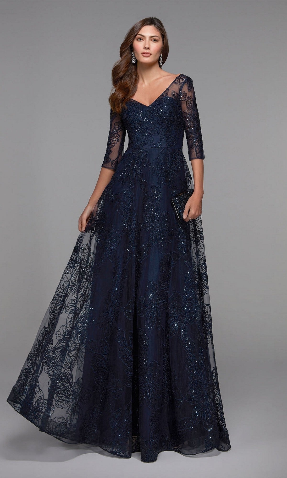 A woman models the Alyce 27494 long navy formal dress, featuring sheer sequin lace sleeves and a V-neckline, standing against a plain gray backdrop.